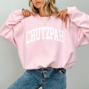 Pu&ograve; includere: Felpa rosa chiaro con la scritta "CHUTZPAH" stampata in bianco sul petto. La felpa ha maniche lunghe e girocollo. La modella indossa jeans blu.