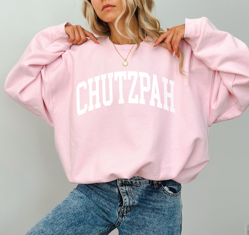 Pu&ograve; includere: Felpa rosa chiaro con la scritta "CHUTZPAH" stampata in bianco sul petto. La felpa ha maniche lunghe e girocollo. La modella indossa jeans blu.