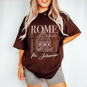 May include: A brown t-shirt with a white graphic design of the Colosseum in Rome, Italy. The text "ROME Italy Family Trip Summer '23 The Johnsons" is printed on the shirt.