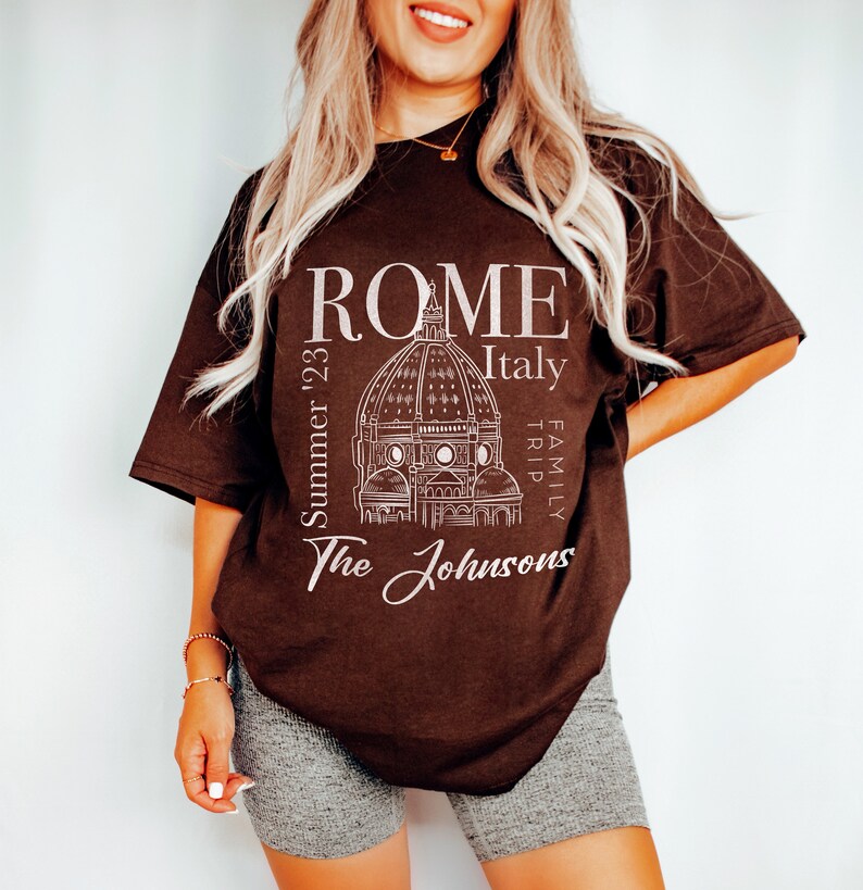 May include: A brown t-shirt with a white graphic design of the Colosseum in Rome, Italy. The text "ROME Italy Family Trip Summer '23 The Johnsons" is printed on the shirt.