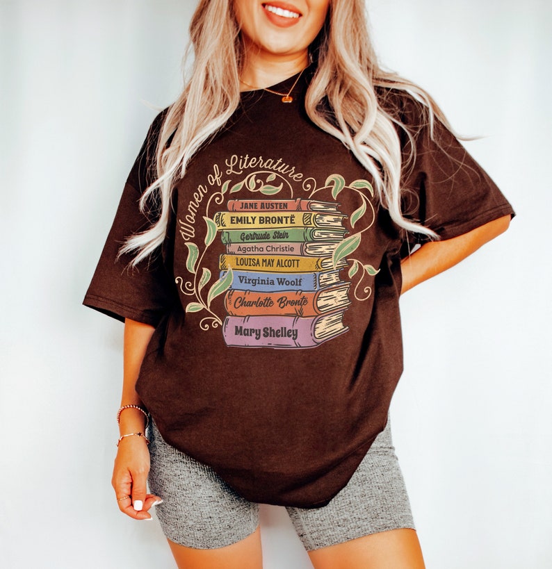May include: A brown t-shirt with a graphic of a stack of books featuring the names of famous women of literature, including Jane Austen, Emily Bronte, Gertrude Stein, Agatha Christie, Louisa May Alcott, Virginia Woolf, Charlotte Bronte, and Mary Shelley. The text "Women of Literature" is written in a circular shape around the books.