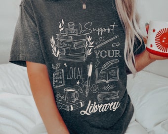 May include: A dark gray t-shirt with a white graphic design featuring a stack of books, a pen, a cup of coffee, and the text "Support Your Local Library".