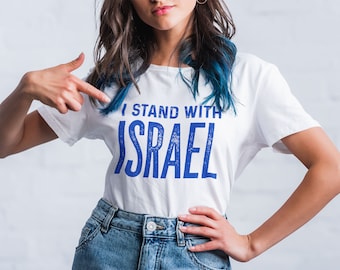May include: A woman wearing a white t-shirt with blue text that reads "I stand with Israel". She is pointing at the shirt with her right hand.