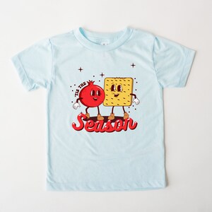 Op de afbeelding: Een lichtblauw t-shirt met een rode granaatappel en een gele cracker met gezichten. De tekst "Tis the Season" staat in rood onder de figuren.