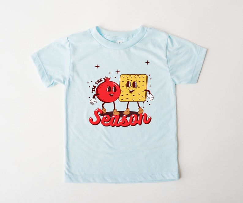Op de afbeelding: Een lichtblauw t-shirt met een rode granaatappel en een gele cracker met gezichten. De tekst "Tis the Season" staat in rood onder de figuren.