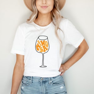 May include: White t-shirt with a graphic of a wine glass with the word "MIMOSA" inside the glass in orange and yellow letters.