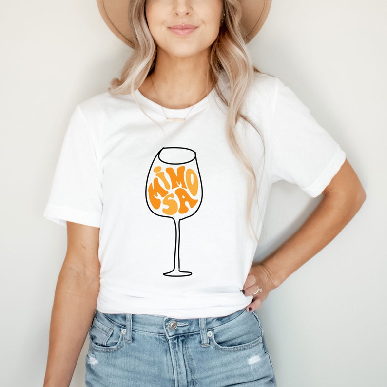 May include: White t-shirt with a graphic of a wine glass with the word "MIMOSA" inside the glass in orange and yellow letters.