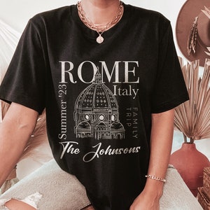 May include: Black t-shirt with a white graphic design of the Colosseum in Rome, Italy. The text "ROME" is above the image, and "Italy" is to the right. The text "Summer '23" is to the left of the image, and "Family Trip" is to the right. The text "The Johnsons" is below the image.