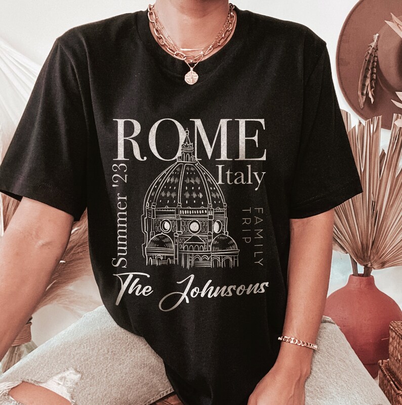 May include: Black t-shirt with a white graphic design of the Colosseum in Rome, Italy. The text "ROME" is above the image, and "Italy" is to the right. The text "Summer '23" is to the left of the image, and "Family Trip" is to the right. The text "The Johnsons" is below the image.