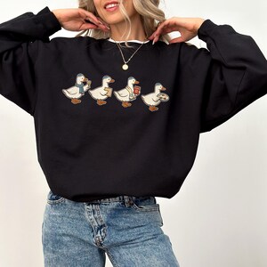 May include: Black sweatshirt featuring a line of four cartoon ducks, each with a different accessory: a scarf, a book, a gift, and a plate of food. The sweatshirt has a white collar and cuffs.