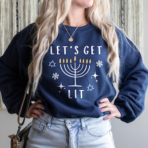 以下が含まれることがあります： 「Let's Get Lit」という文字とメノラーのグラフィックがプリントされたネイビーブルーのスウェットシャツ。メノラーは金色で、炎は黄色です。