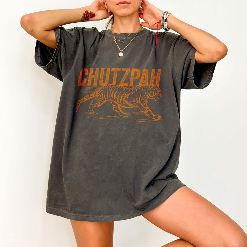 May include: A dark gray oversized t-shirt with an orange graphic design. The design features the word 'CHUTZPAH' in bold letters above an orange tiger illustration. The shirt has short sleeves and a relaxed fit, suggesting a casual style.