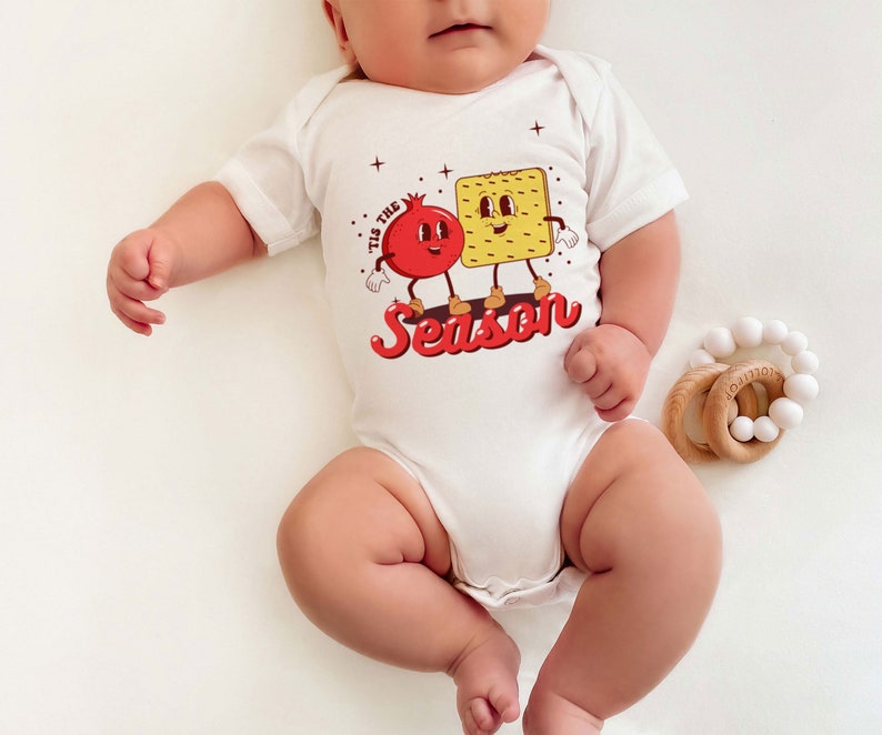 Op de afbeelding: Witte baby romper met een rode granaatappel en een gele cracker met gezichten. De tekst 'Tis the Season' is gedrukt in rode en gele letters.