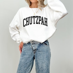 Pu&ograve; includere: Felpa bianca con la scritta "CHUTZPAH" stampata a caratteri neri in grassetto sul petto. La felpa &egrave; a maniche lunghe e ha un girocollo. La persona indossa jeans azzurri.