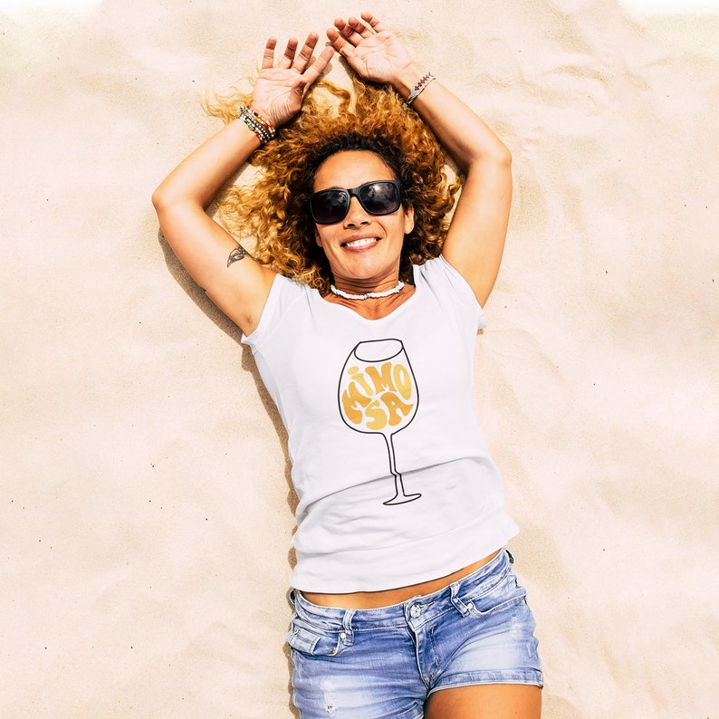 May include: A woman wearing a white t-shirt with a gold graphic of a wine glass that says "Mimosa" on a sandy beach. She is wearing sunglasses and has her arms raised above her head.