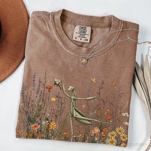 Puede incluir: Una camiseta marrón con un estampado gráfico de una mantis religiosa verde entre flores silvestres y lavanda. La camiseta presenta una etiqueta de la marca Comfort Colors en el cuello interior y un collar de cadena plateada.