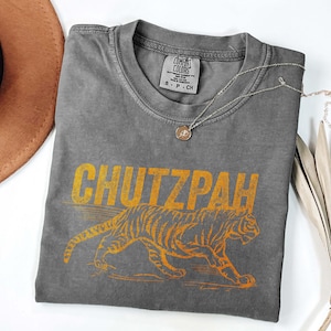 May include: A gray Comfort Colors t-shirt with the word "CHUTZPAH" in yellow block letters above a yellow tiger illustration. The shirt has a crew neck and a small tag on the collar. A silver necklace with a pendant is draped across the shirt.