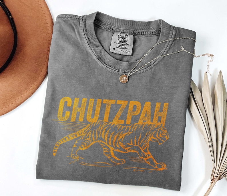 May include: A gray Comfort Colors t-shirt with the word "CHUTZPAH" in yellow block letters above a yellow tiger illustration. The shirt has a crew neck and a small tag on the collar. A silver necklace with a pendant is draped across the shirt.