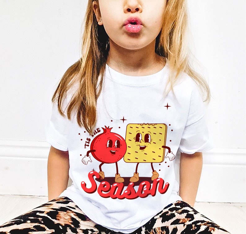 Op de afbeelding: Wit T-shirt met een rode granaatappel en een gele cracker met gezichten. De tekst "Tis the Season" staat in rood onder de figuren.