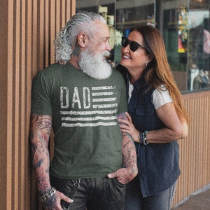 May include: A couple standing near a building. The man is wearing a green t-shirt with the word "DAD" and a distressed American flag design. The woman is wearing a white shirt and a denim vest.