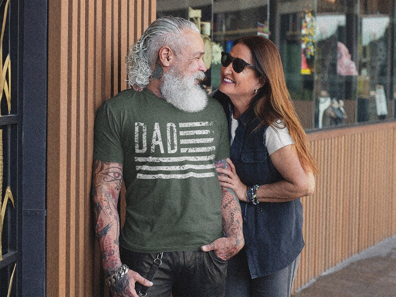 May include: A couple standing near a building. The man is wearing a green t-shirt with the word "DAD" and a distressed American flag design. The woman is wearing a white shirt and a denim vest.
