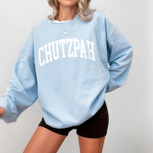 Pu&ograve; includere: Una felpa azzurra con la scritta "CHUTZPAH" in bianco, a lettere arcuate. La felpa ha maniche lunghe e girocollo. La modella indossa pantaloncini neri e una collana d'argento.