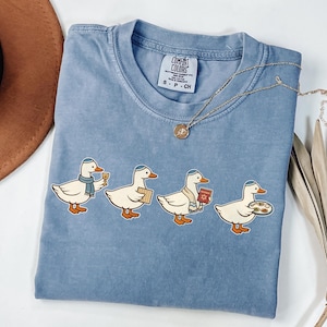 May include: A light blue Comfort Colors t-shirt featuring four cartoon ducks, each wearing a yarmulke and holding items associated with Passover. The ducks are holding a glass of wine, matzah, a Haggadah, and a seder plate.