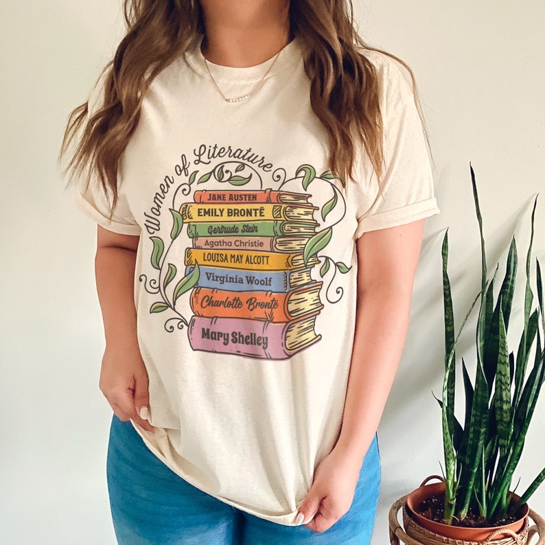 May include: A cream colored t-shirt with a graphic of a stack of books with the names of famous women authors printed on the spines. The text "Women of Literature" is printed above the books.