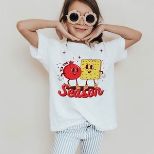 Op de afbeelding: Wit T-shirt met een rode granaatappel en een gele cracker met gezichten. De tekst "Tis the Season" is geschreven in rode en witte letters.