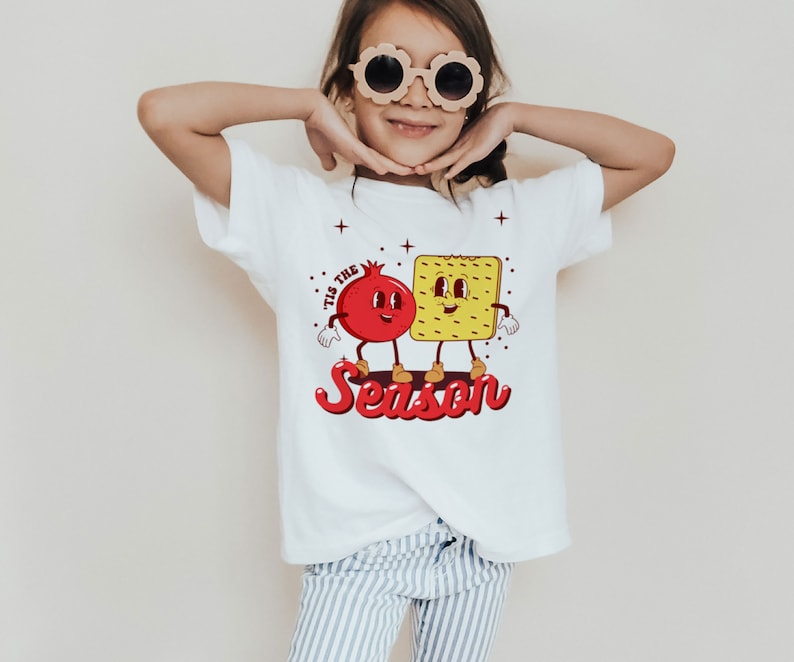 Op de afbeelding: Wit T-shirt met een rode granaatappel en een gele cracker met gezichten. De tekst "Tis the Season" is geschreven in rode en witte letters.