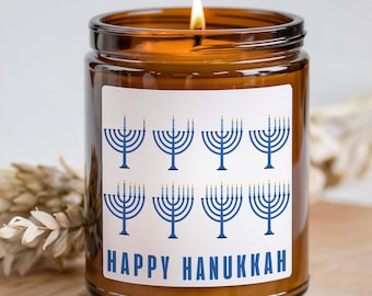 May include: A lit Hanukkah candle in a brown glass jar. The jar has a white label with blue menorahs and the words "Happy Hanukkah" in blue. The candle is on a wooden surface.