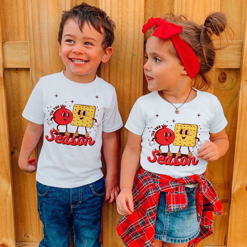 Op de afbeelding: Twee kinderen dragen witte T-shirts met een rode granaatappel en een gele cracker afbeelding. De shirts zeggen "Tis the Season". Het meisje draagt een rood hoofdband en een rood-zwarte geruite rok.