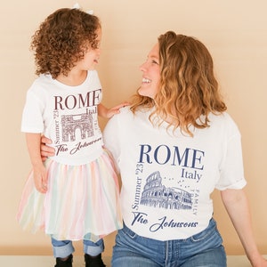 May include: A white t-shirt with a graphic of the Colosseum in Rome, Italy. The text "Summer '23" and "The Johnsons" are printed on the shirt. A matching t-shirt with a graphic of the Arch of Constantine is worn by a child.
