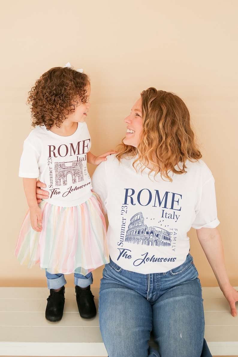 May include: A white t-shirt with a graphic of the Colosseum in Rome, Italy. The text "Summer '23" and "The Johnsons" are printed on the shirt. A matching t-shirt with a graphic of the Arch of Constantine is worn by a child.