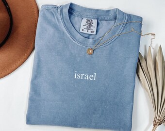 May include: A blue Comfort Colors t-shirt with the word "israel" in white lettering. A gold necklace with a pendant rests on the shirt. A brown hat and dried plant are in the background.