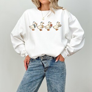 May include: White sweatshirt featuring a whimsical design of four cartoon ducks, each carrying different items like a coffee cup, package, and pizza. The ducks are wearing hats and scarves. The sweatshirt is paired with blue jeans.