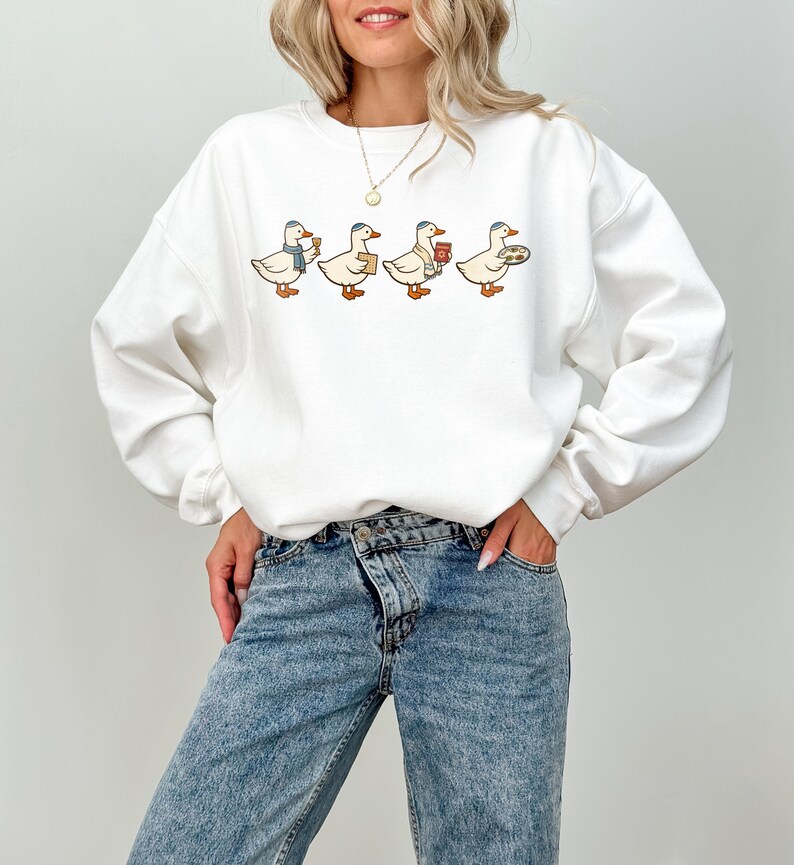 May include: White sweatshirt featuring a whimsical design of four cartoon ducks, each carrying different items like a coffee cup, package, and pizza. The ducks are wearing hats and scarves. The sweatshirt is paired with blue jeans.