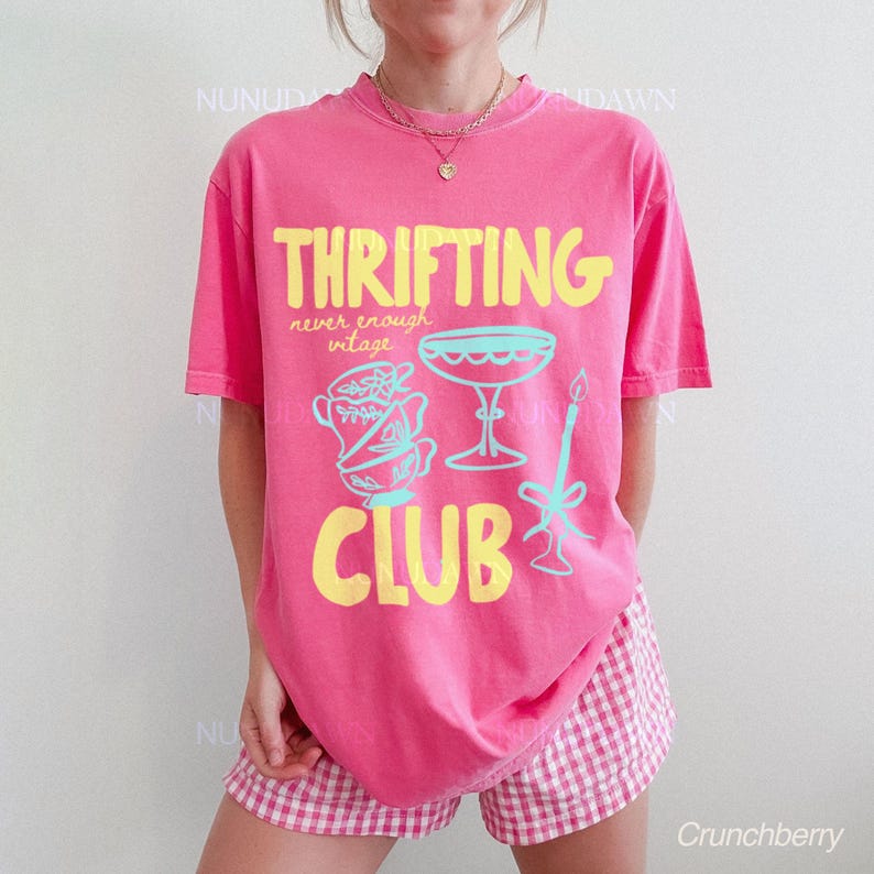 以下が含まれることがあります： ピンク色のTシャツ。黄色とターコイズ色のグラフィックで「THRIFTING CLUB」の文字。ティーカップ、シャンパングラス、キャンドルのヴィンテージ風デザイン。モデルはピンクと白のチェック柄のショートパンツを着用。