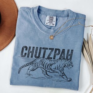 May include: A blue Comfort Colors t-shirt with the word "CHUTZPAH" above a tiger illustration. A gold necklace with a pendant is draped across the shirt. A brown hat and dried plant are in the background.