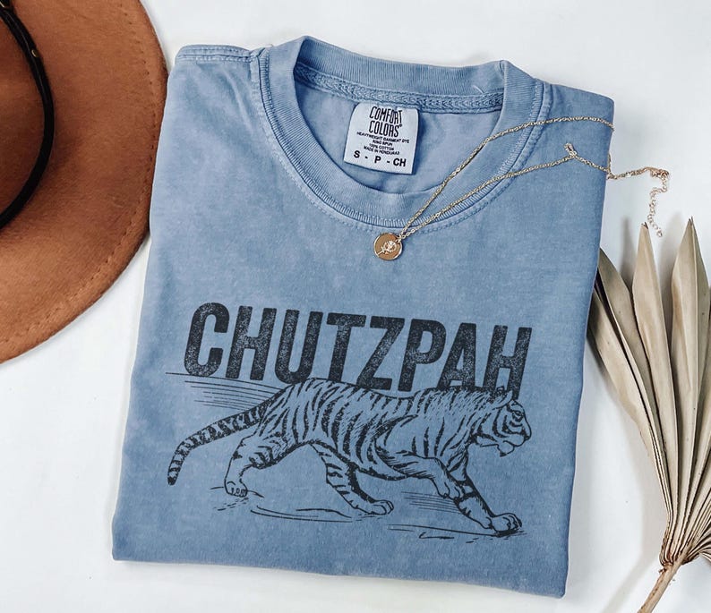 May include: A blue Comfort Colors t-shirt with the word "CHUTZPAH" above a tiger illustration. A gold necklace with a pendant is draped across the shirt. A brown hat and dried plant are in the background.
