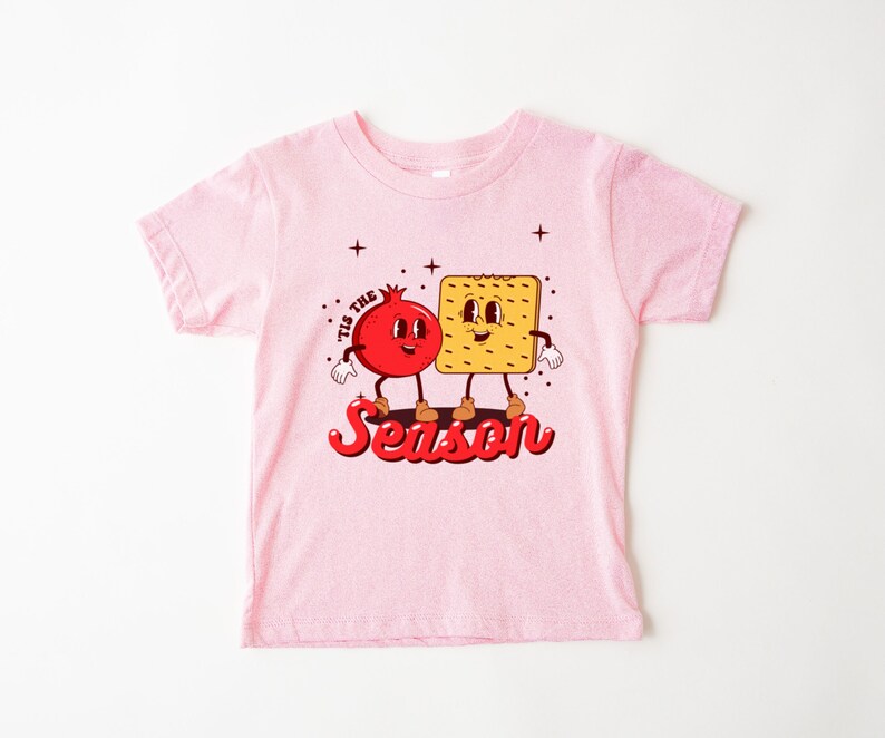 Op de afbeelding: Roze peuter T-shirt met een cartoon granaatappel en cracker met de tekst "Tis the Season".
