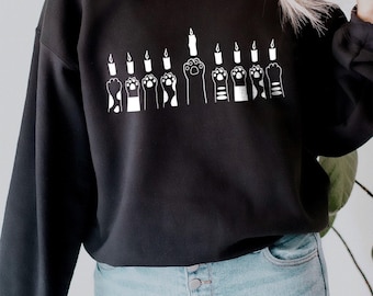 May include: Black sweatshirt featuring a white graphic of paw prints holding candles. The design is a playful take on the traditional menorah.