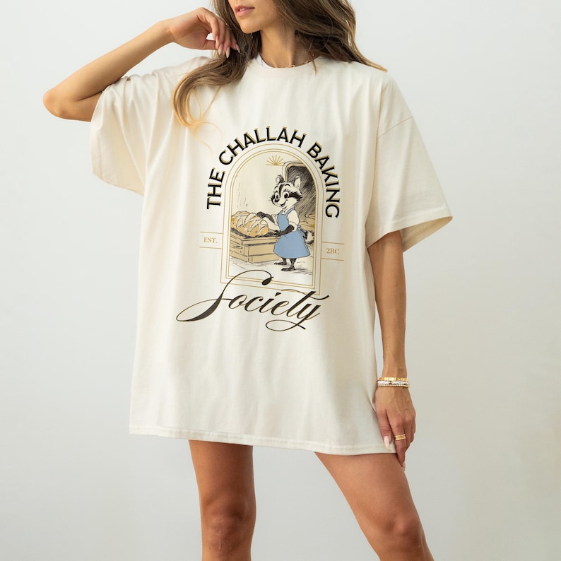 K&ouml;nnte beinhalten: Cremefarbenes T-Shirt mit einem Waschb&auml;r-B&auml;cker-Motiv. Der Waschb&auml;r tr&auml;gt eine blaue Sch&uuml;rze und h&auml;lt Brot. Der Text "THE CHALLAH BAKING Society" und "EST. 2010" sind ebenfalls auf dem Shirt abgebildet. Das Shirt ist oversized.