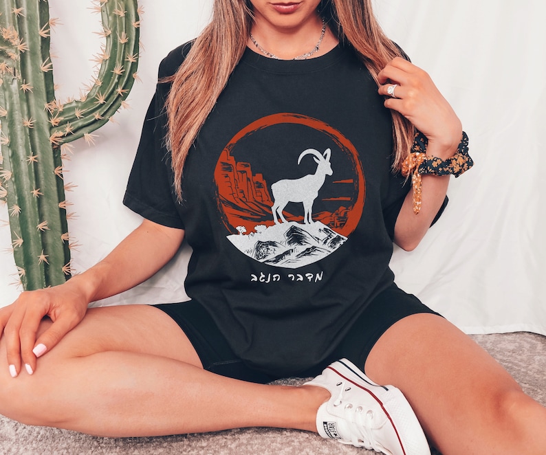 May include: Black t-shirt featuring a white mountain goat graphic inside a red and orange circular design. The design includes a mountain range and text in Hebrew. The shirt is a casual wear item.