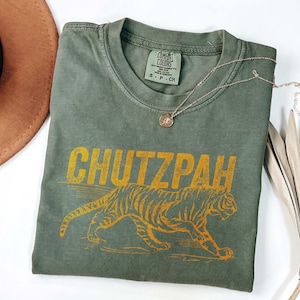 May include: Olive green t-shirt with the word "CHUTZPAH" in yellow block letters above a yellow tiger illustration. The shirt has a Comfort Colors tag and a silver necklace with a small pendant.