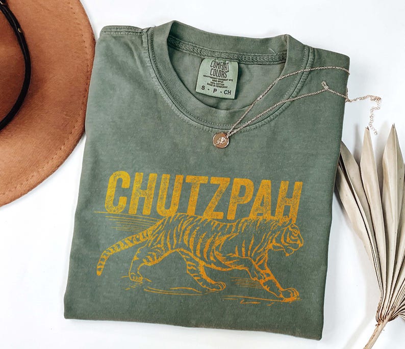 May include: Olive green t-shirt with the word "CHUTZPAH" in yellow block letters above a yellow tiger illustration. The shirt has a Comfort Colors tag and a silver necklace with a small pendant.