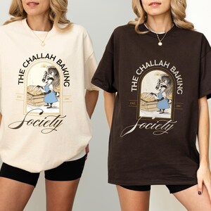 K&ouml;nnte beinhalten: Zwei T-Shirts, eines in Creme und eines in Braun, mit einer Grafik eines B&auml;ckerracoon und dem Text "The Challah Baking Society". Das Design beinhaltet ein bogenf&ouml;rmiges Fenster und das Wort "Society" in Schreibschrift. Die Shirts sind kurz&auml;rmelig.