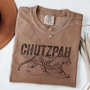 May include: A brown t-shirt with the word "CHUTZPAH" printed in black, along with a tiger illustration. The shirt has a crew neck and short sleeves. A silver necklace with a small pendant rests on the shirt.