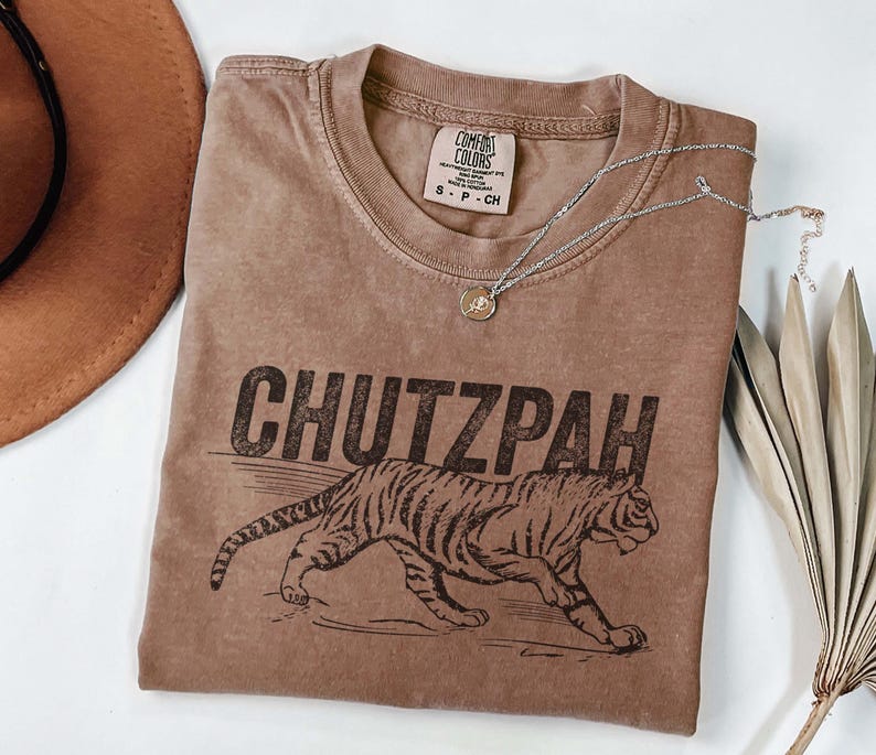 May include: A brown t-shirt with the word "CHUTZPAH" printed in black, along with a tiger illustration. The shirt has a crew neck and short sleeves. A silver necklace with a small pendant rests on the shirt.