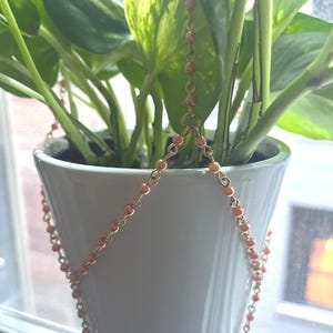May include: A white ceramic planter with a trailing plant and a decorative chain plant hanger. The chain is made of small, peach-colored beads strung on a gold-colored chain. The plant has green and yellow variegated leaves.